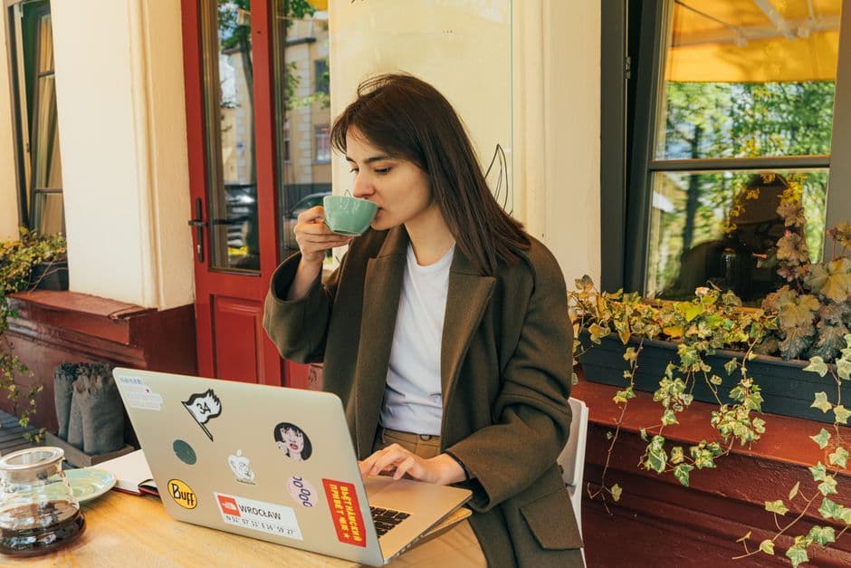 Young woman drinking coffee and working on laptop in outdoor cafe setting.